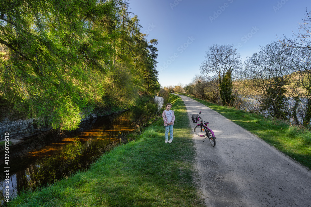 A girl and her bike rests peacefully by a calm and serene canal, enjoys the lush greenery, gentle breeze, and warm sunlight in this idyllic landscape, Bohernabreena Reservoir, Dublin, Ireland