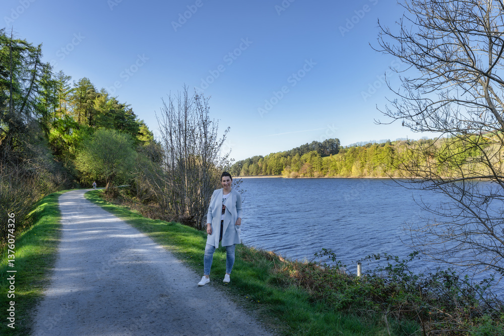 A woman takes great pleasure in a serene moment by the side of a peaceful lake, encompassed by vibrant green trees and an expansive clear blue sky overhead, Bohernabreena Reservoir, Dublin, Ireland