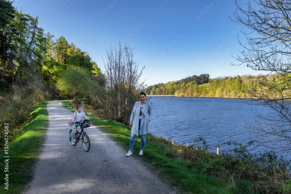 On a beautiful sunny day, a mother and her child joyfully ride a bicycle together along a picturesque pathway that runs beside a river surrounded by nature, Bohernabreena Reservoir, Dublin, Ireland