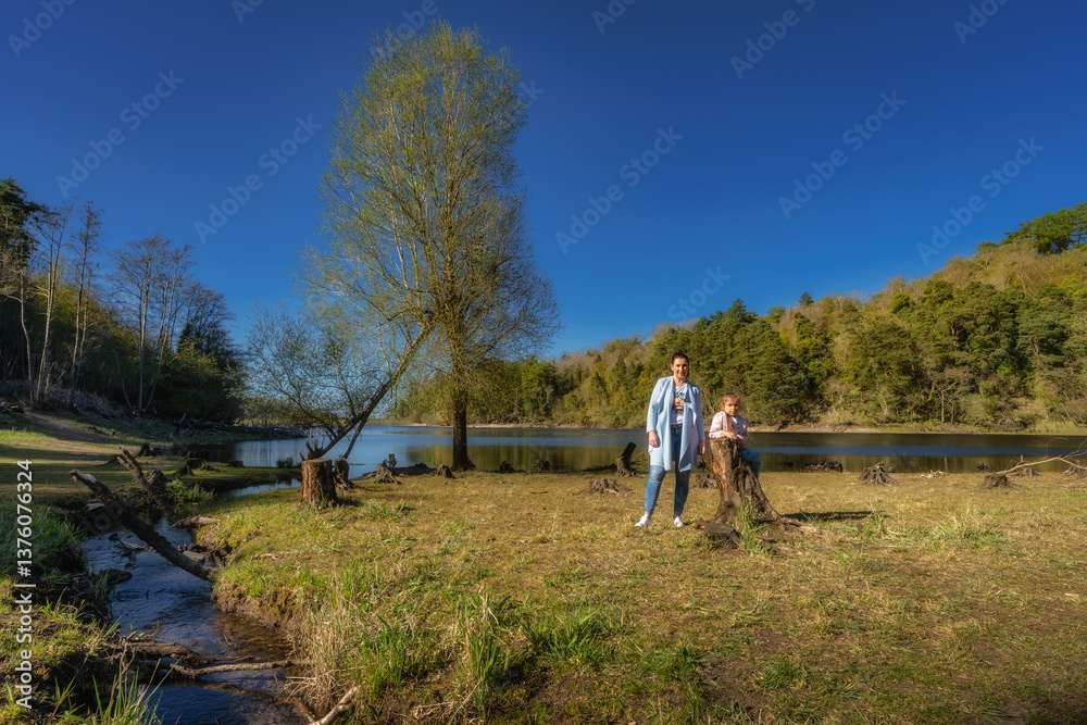 A loving mother and her joyful child stand by a tranquil lake, surrounded by lush greenery and vibrant flowers, creating a picturesque scene for bonding, Bohernabreena Reservoir, Dublin, Ireland