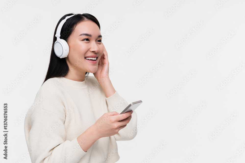 Smiling Asian young woman girl female student in white headphones earphones looking aside holds mobile smart phone listening music choosing song podcast isolated on white background. Smiling Asian young woman girl female student in white headphones earphones looking aside holds mobile smart phone listening music choosing song podcast isolated on white background.