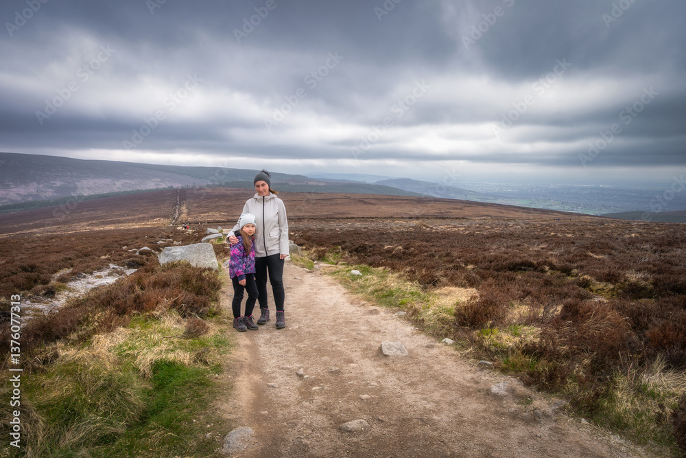 A loving mother and her daughter stand side by side on a breathtaking hiking trail, surrounded by lush greenery and a dramatic, cloudy sky that adds to their adventure, Wicklow, Dublin, Ireland