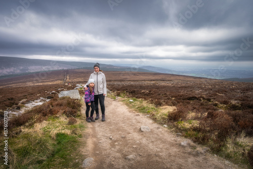 Obraz na płótnie A loving mother and her daughter stand side by side on a breathtaking hiking trail, surrounded by lush greenery and a dramatic, cloudy sky that adds to their adventure, Wicklow, Dublin, Ireland w salonie