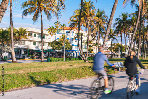 Obraz na płótnie A vibrant street scene in Miami Beach with palm trees, colorful Art Deco-style buildings, and motion blurred cyclists along a sunny beachfront promenade w salonie