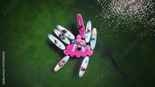 Aerial top-down view of a group practicing yoga on a floating SUP island in the middle of a serene lake. Calm water reflects the sky, creating a peaceful, harmonious scene.
