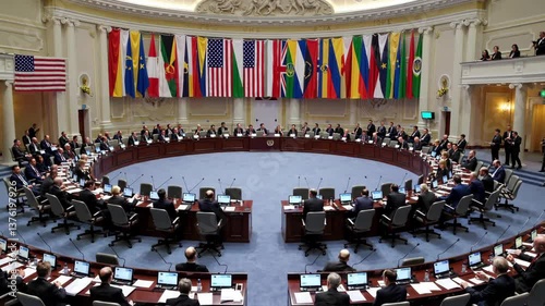 High angle view of a large group of international delegates and diplomats engaging in discussions at a round table, adorned with flags from various nations, during a world peace summit