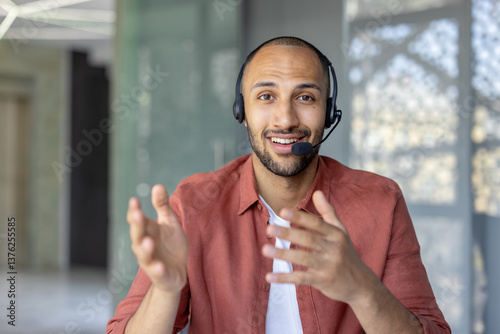 Webcam view, support worker with headset looking into camera, smiling, talking and consulting clients remotely. Man at workplace inside office
