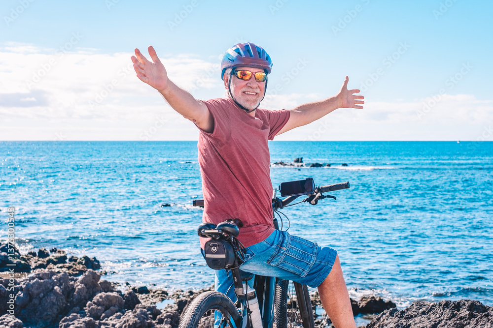 Rear view of active senior man with outstretched arms enjoying a sunny day at the beach with the electric bicycle. Authentic retirement living and healthy lifestyle concept.