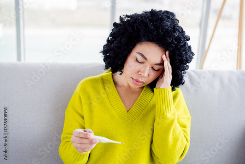 Young woman feeling unwell sitting on a comfortable sofa and checking her temperature with a thermometer