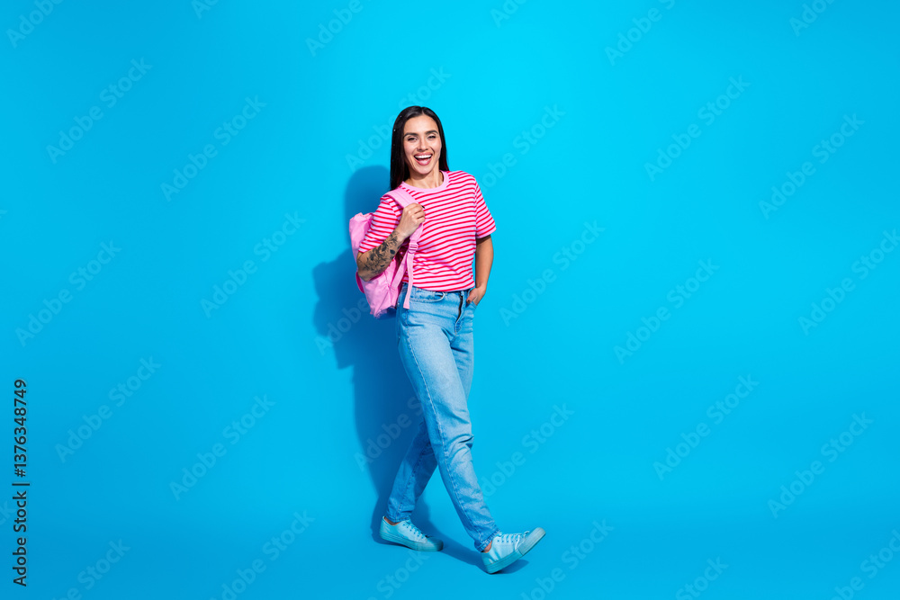 Naklejka premium Stylish young woman wearing a pink striped t-shirt, jeans, and backpack in front of a vibrant blue background exudes charm and confidence.