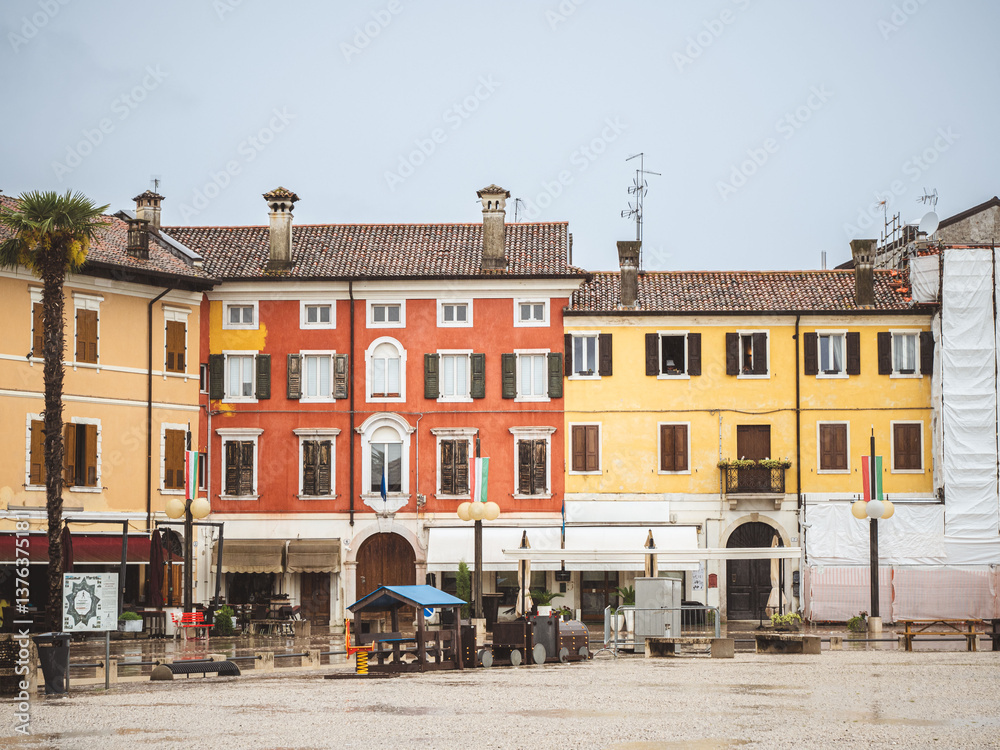 Obraz premium A rainy day in the historic town square of Palmanova, Italy, featuring old buildings with colorful facades, statues, and palm trees under a cloudy sky.