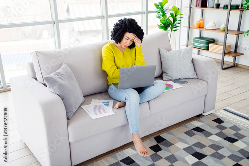 Young woman relaxing on a sofa in a modern living room, using a laptop and working with documents during a casual and comfortable day.