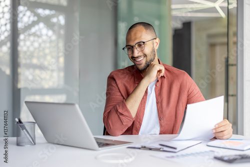 A smiling businessman reviews documents at his desk with a laptop nearby, appearing focused and content in the office.