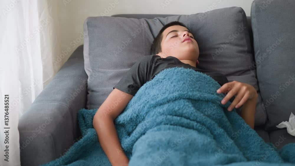 Uncomfortable child sleeping covered in blanket adjusting on couch for comfort with tissue nearby, showing signs of discomfort during rest in peaceful indoor setting