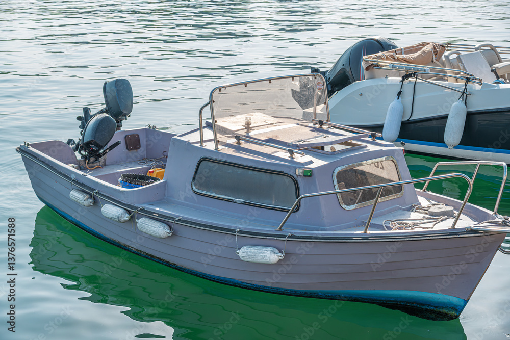 A small, weathered motorboat with a cabin is moored in a marina, equipped with an outboard motor, protective fenders, and fishing gear, floating on calm, sunlit water.