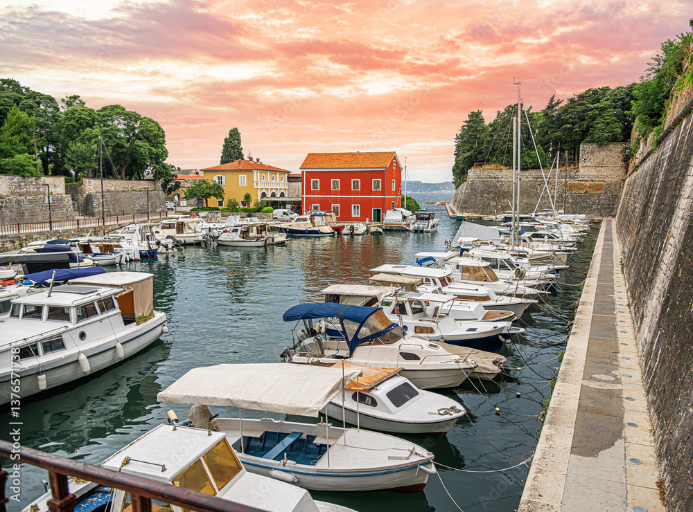 A stunning harbor at sunset, featuring moored boats, colorful historic buildings, and ancient stone walls, creating a picturesque Mediterranean coastal scene.