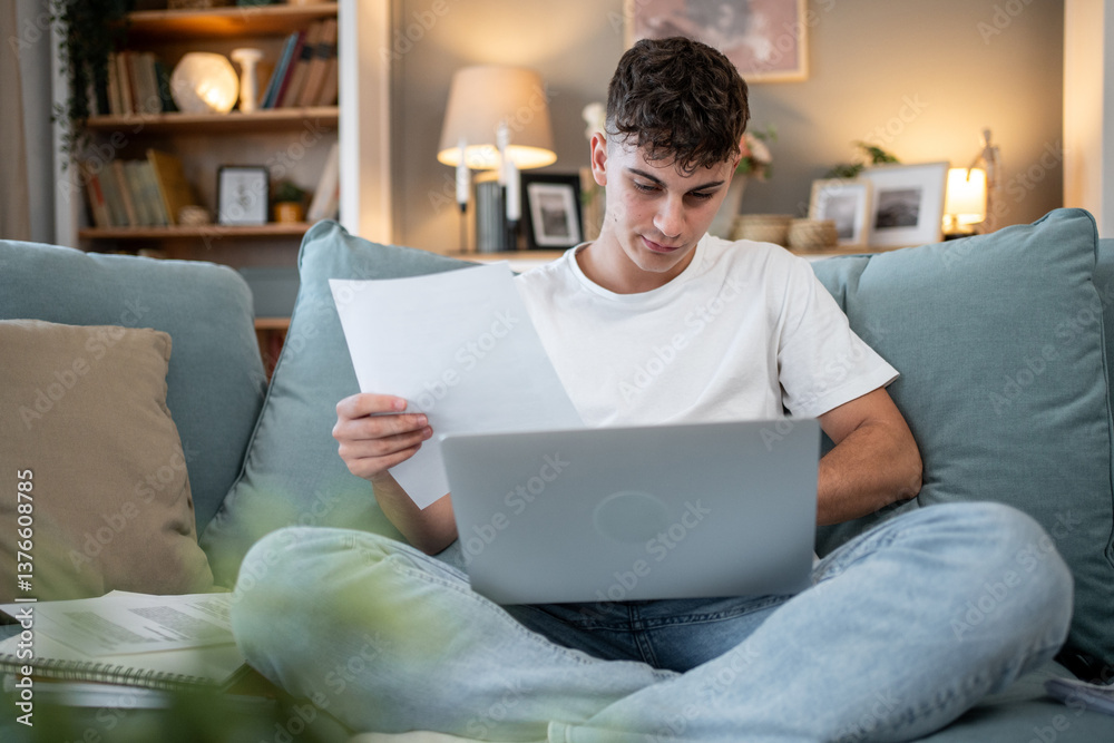 Focused young man working from home sofa using laptop and reading documents Focused young man working from home sofa using laptop and reading documents