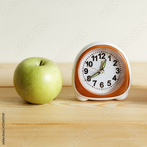 A green apple and a white small alarm clock on a light background.