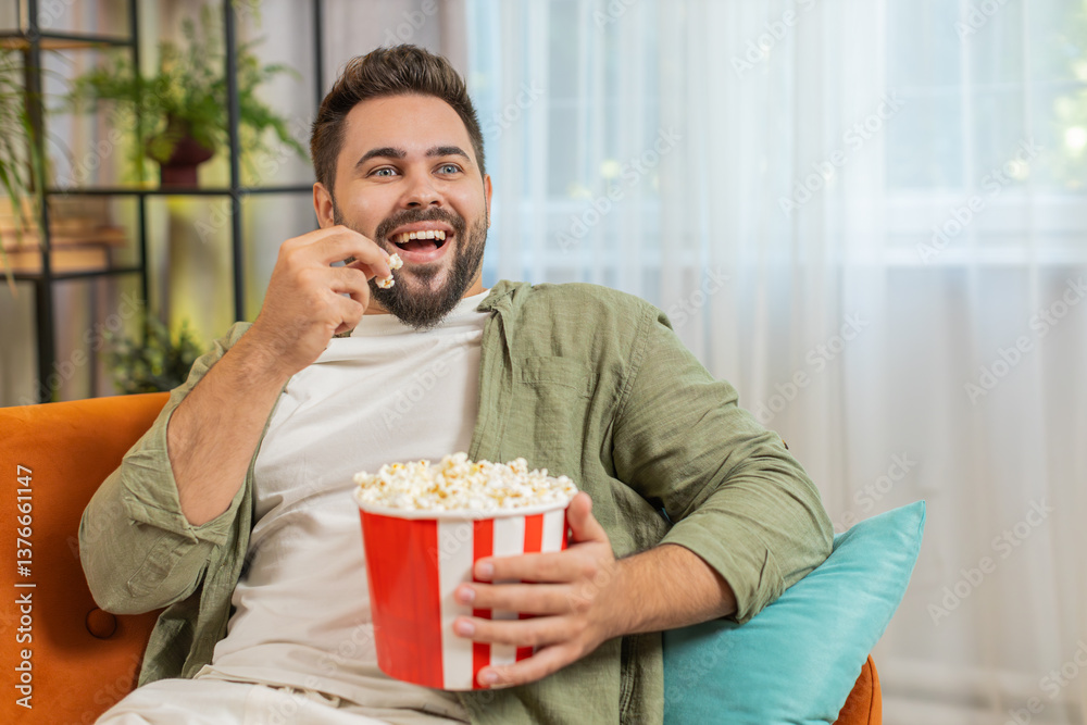 custom made wallpaper toronto digitalRelaxed Caucasian young man eating popcorn and watching movie sitting on sofa in living room at home. Happy guy with brown hair in casual clothes enjoying suspense film during weekend in apartment.
