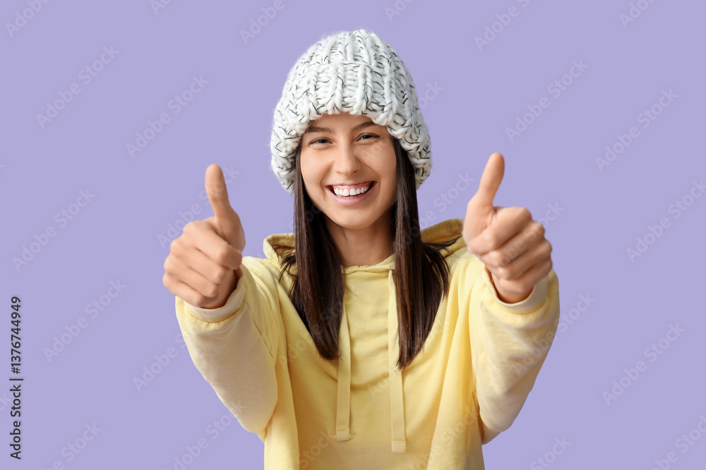 Beautiful young woman in stylish knitted hat showing thumb-up gesture on lilac background Beautiful young woman in stylish knitted hat showing thumb-up gesture on lilac background