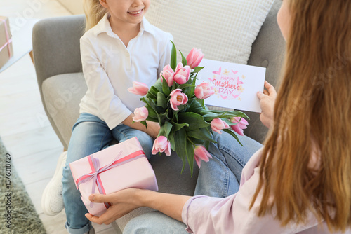 Cute little girl greeting her mom for Mother's Day with tulips, postcard and gift box on sofa in living room, closeup
