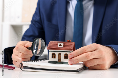 Real estate agent with house figure, notepad and magnifying glass at white table in office, closeup. Home appraisal