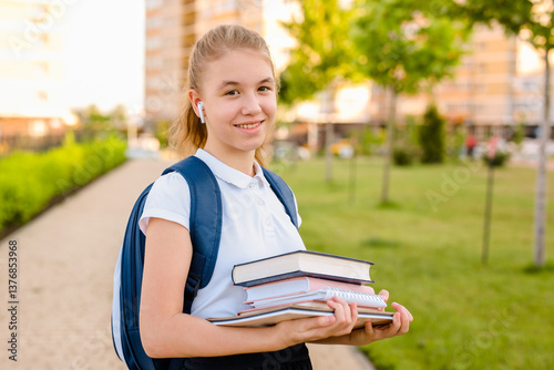 Portrait of smiling teenage schoolgirl with books standing in park near residential buildings