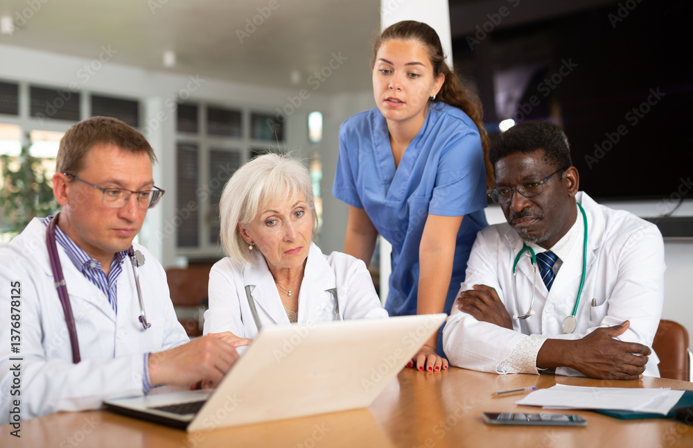 Group of different people doctors in medical uniforms looking attentively at laptop screen on table in office Group of different people doctors in medical uniforms looking attentively at laptop screen on table in office