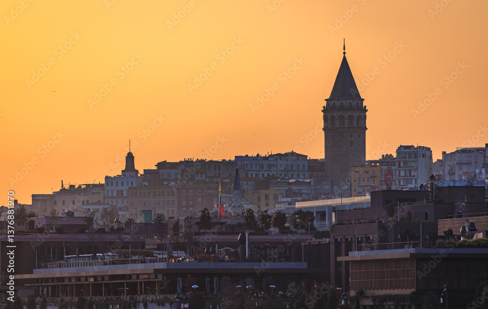 Naklejka premium Galata Tower silhouette stands against golden sunset sky, Istanbul cityscape layered with buildings and rooftops, urban skyline in Turkey at dusk