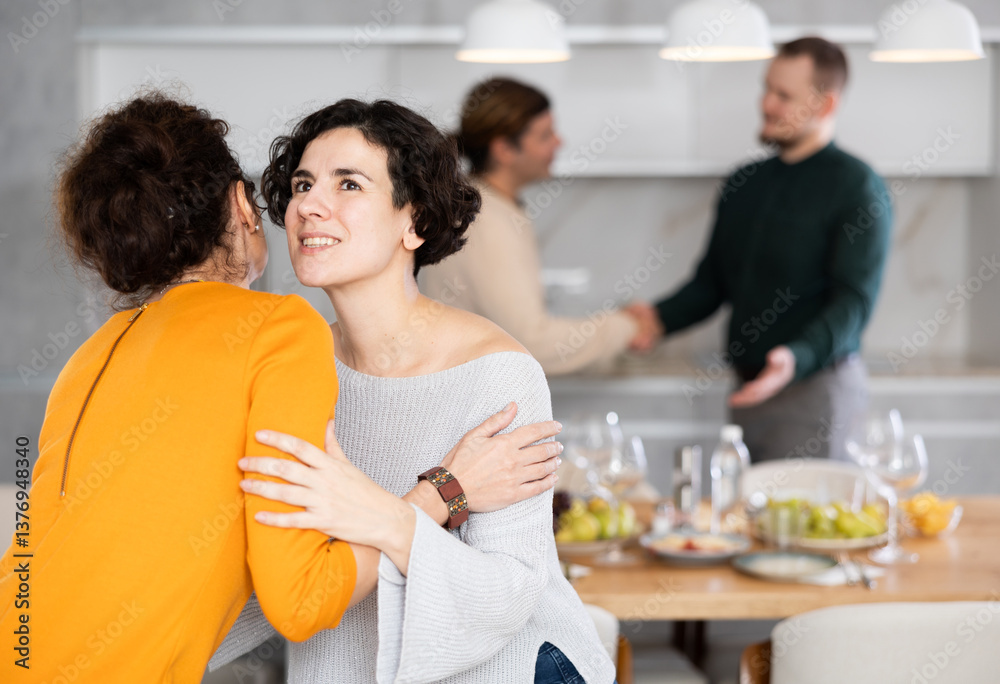Fototapeta premium Women hug each other and kiss on the cheek while greeting each other before a festive dinner. Men shaking hands in the background