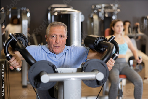 Portrait of concentrated sporty aged man performing lateral raises of arms on deltoid muscle training machine in gym. Concept of sports and fitness for elderly