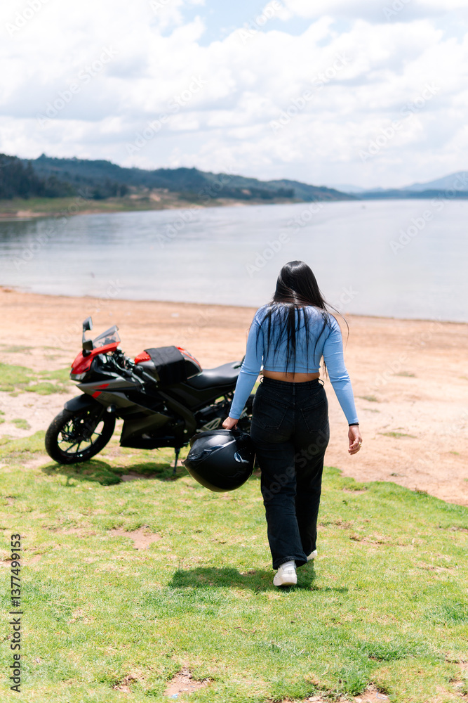 Obraz premium Young woman biker is walking away from her motorcycle parked near a lake, carrying her helmet