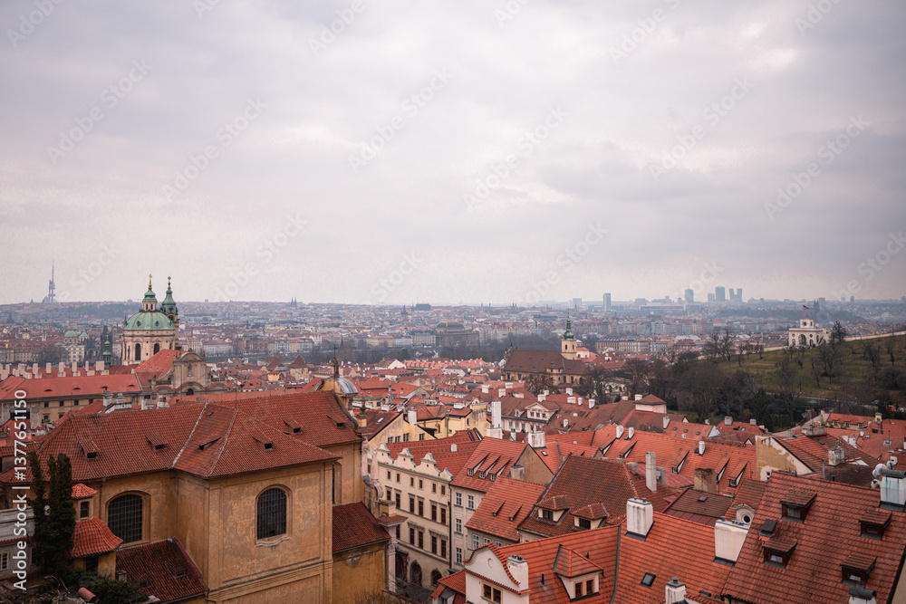 Obraz premium Panoramic view of Prague with red rooftops, St. Nicholas Church, Petřín Hill, and the U.S. Embassy pavilion, under an overcast sky with the city skyline in the distance.