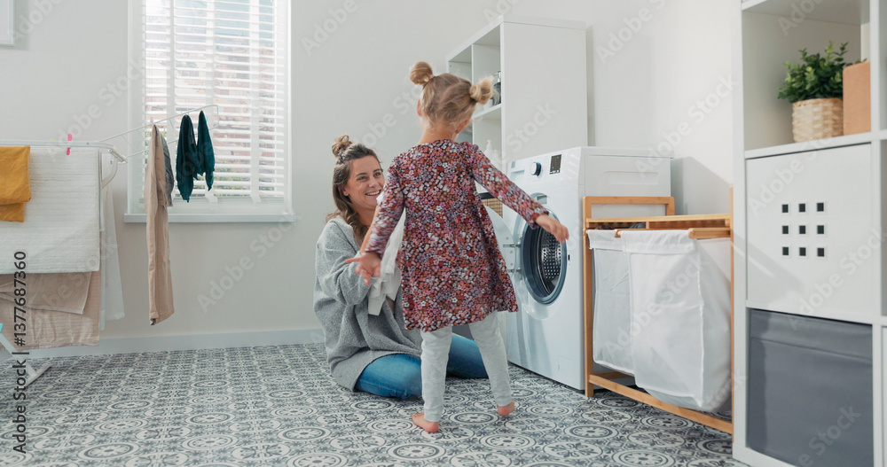 The mother kneels by the washing machine with her little daughter beside her. The girl offers garments from the basket as they smile during chores. The mother kneels by the washing machine with her little daughter beside her. The girl offers garments from the basket as they smile during chores.