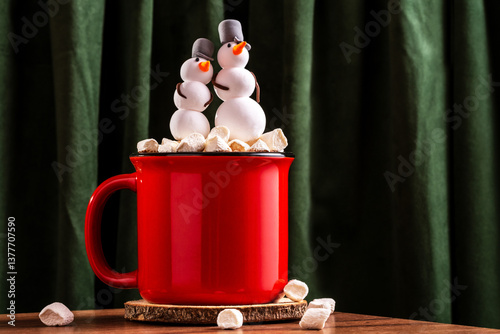 A red mug with a hot drink and marshmallows in form of snowman  on wooden table on background of green velvet curtains