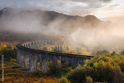 Obraz na płótnie The morning train steams across the Glenfinnan Viaduct, its plume rising against the Highland mist. Golden light touches the hills, creating a scene of timeless beauty in Scotland’s heart. w salonie
