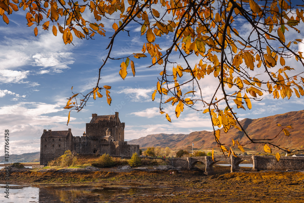 Eilean Donan Castle stands majestically amid autumn hues, its reflection shimmering in calm waters. Misty Highlands and golden trees create a scene of timeless beauty.