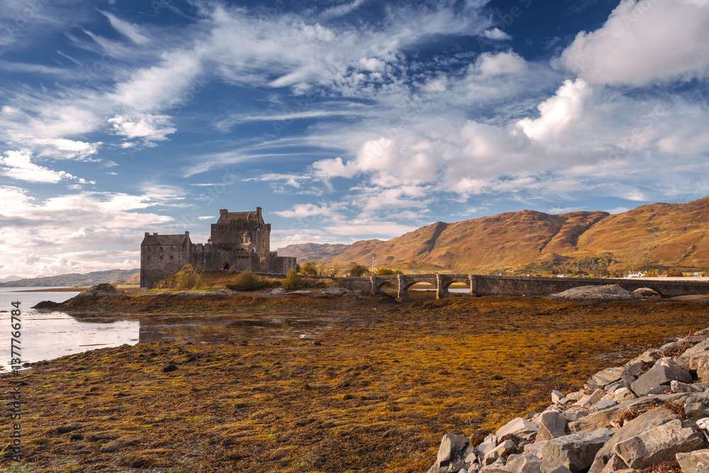 Eilean Donan Castle stands majestically amid autumn hues, its reflection shimmering in calm waters. Misty Highlands and golden trees create a scene of timeless beauty.