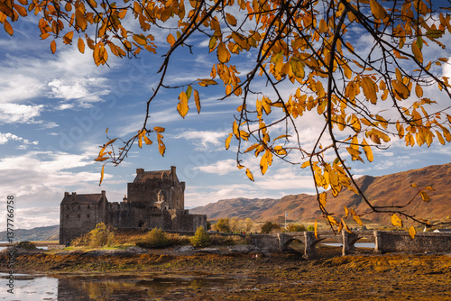 Obraz na płótnie Eilean Donan Castle stands majestically amid autumn hues, its reflection shimmering in calm waters. Misty Highlands and golden trees create a scene of timeless beauty. w salonie
