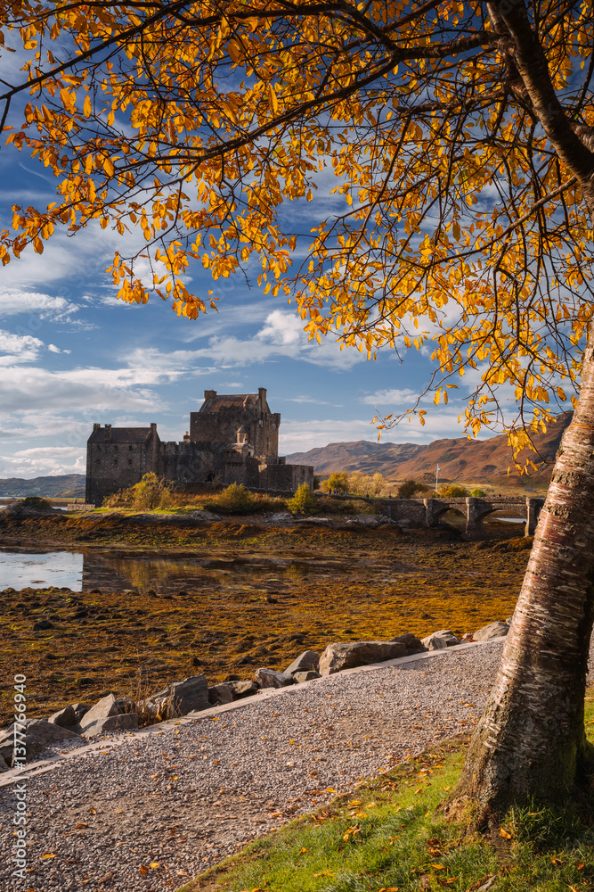 Eilean Donan Castle stands majestically amid autumn hues, its reflection shimmering in calm waters. Misty Highlands and golden trees create a scene of timeless beauty.