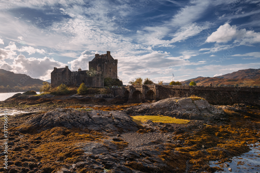 Eilean Donan Castle stands majestically amid autumn hues, its reflection shimmering in calm waters. Misty Highlands and golden trees create a scene of timeless beauty.
