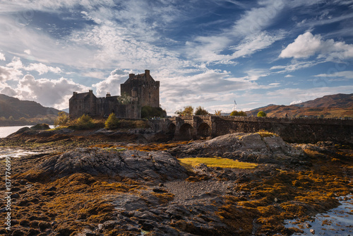 Obraz na płótnie Eilean Donan Castle stands majestically amid autumn hues, its reflection shimmering in calm waters. Misty Highlands and golden trees create a scene of timeless beauty. w salonie