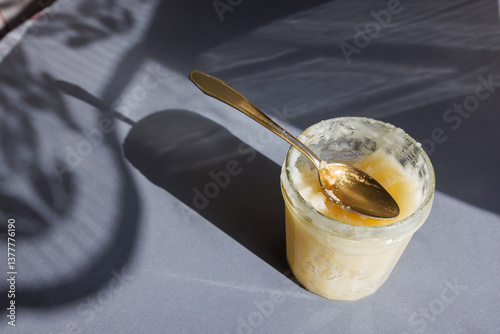 Jar of raw crystallized honey with spoon on grey background with sharp shadows. Organic food.