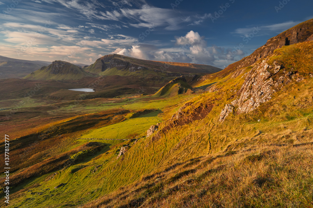 Autumn light bathes the Quiraing on Skye, casting long shadows over its dramatic cliffs and rolling hills. The landscape glows with golden hues, a breathtaking Highland scene.