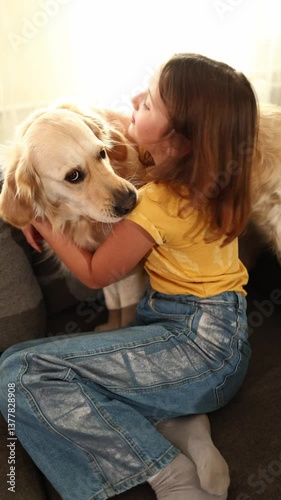 A young girl with brown hair smiles as she hugs a golden retriever standing on a couch. The well-lit living room adds warmth to their joyful moment together.