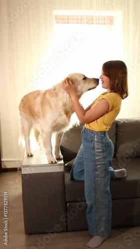 A young girl with brown hair smiles as she hugs a golden retriever standing on a couch. The well-lit living room adds warmth to their joyful moment together.