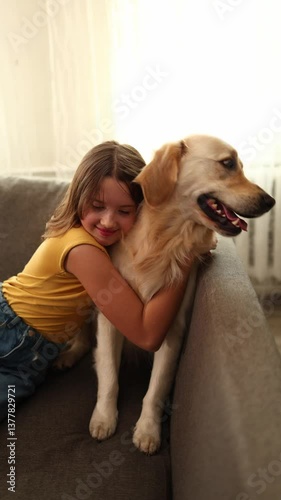 A young girl with brown hair smiles as she hugs a golden retriever sittting on a couch. The well-lit living room adds warmth to their joyful moment together.