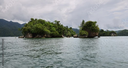 Dramatic limestone islets covered in lush tropical vegetation rise from the calm waters of Salat Rabia, West Papua, framed by misty mountains and a moody, overcast sky.
