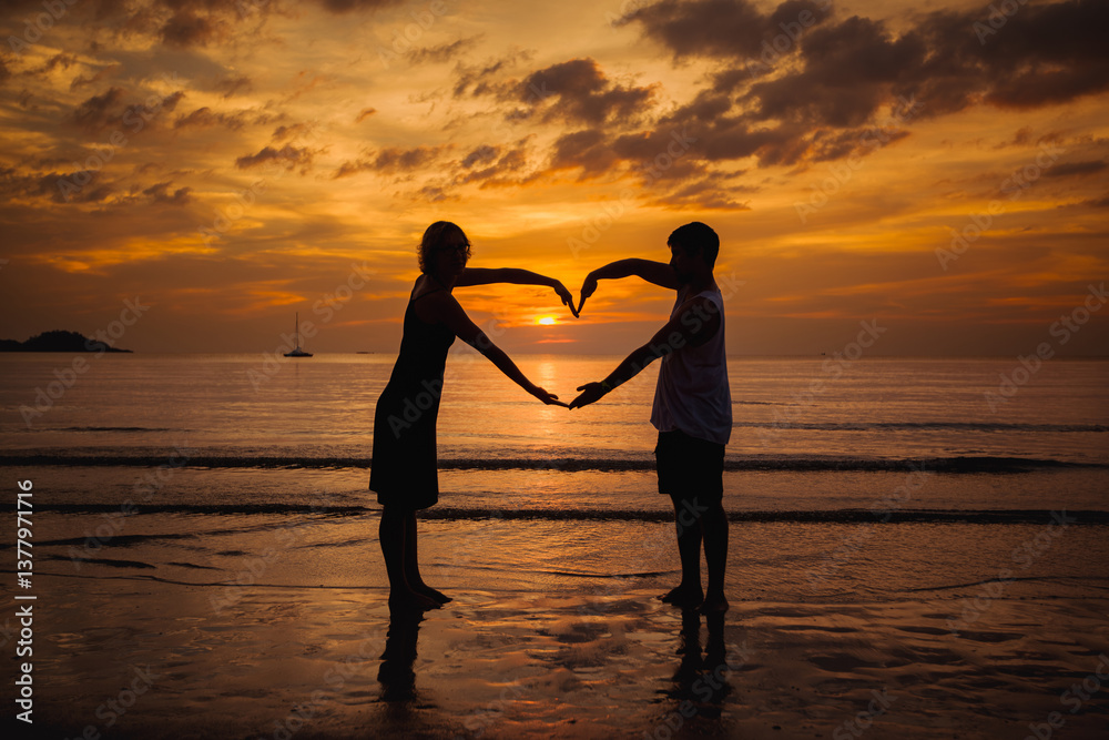 The silhouette of a couple on a seaside beach at twilight, forming a heart with their hands against the backdrop of the setting sun.