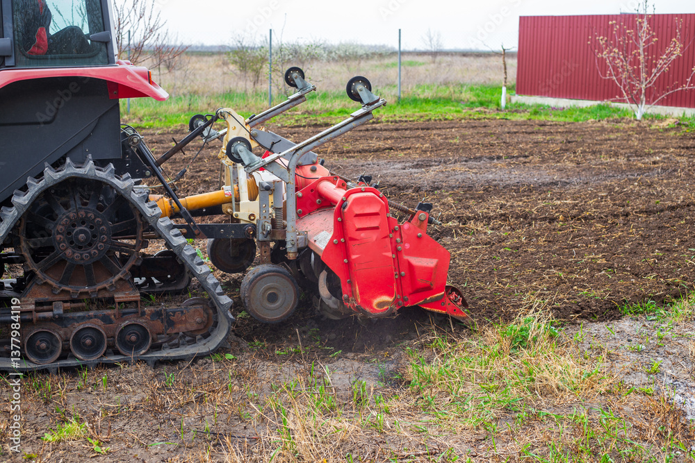 Obraz premium A tractor equipped with a red rotary cultivator actively prepares the soil in a rural field. The equipment loosens the earth, preparing it for planting and cultivation